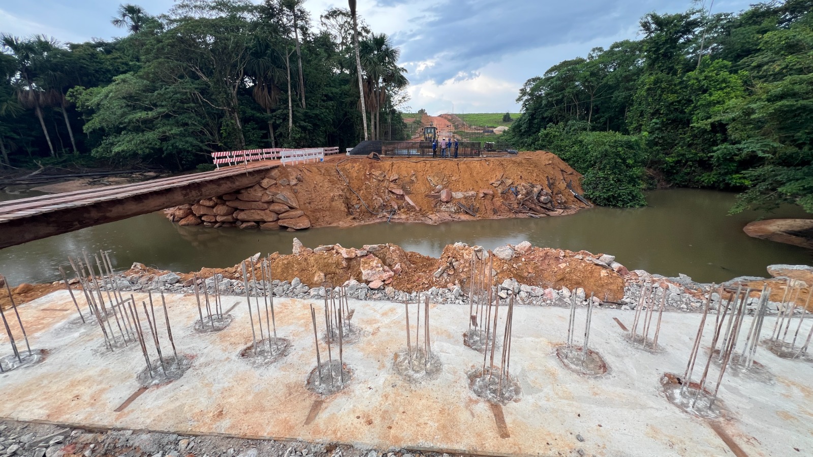 Obra da nova ponte de concreto sobre o Rio Cujubim, na RO-205, com armações de ferro na laje e trabalhadores ao fundo